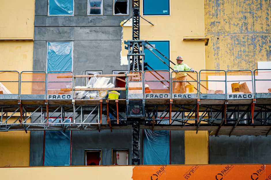 Low angle of male constructor in hardhat standing on crane platform at building wall under construction